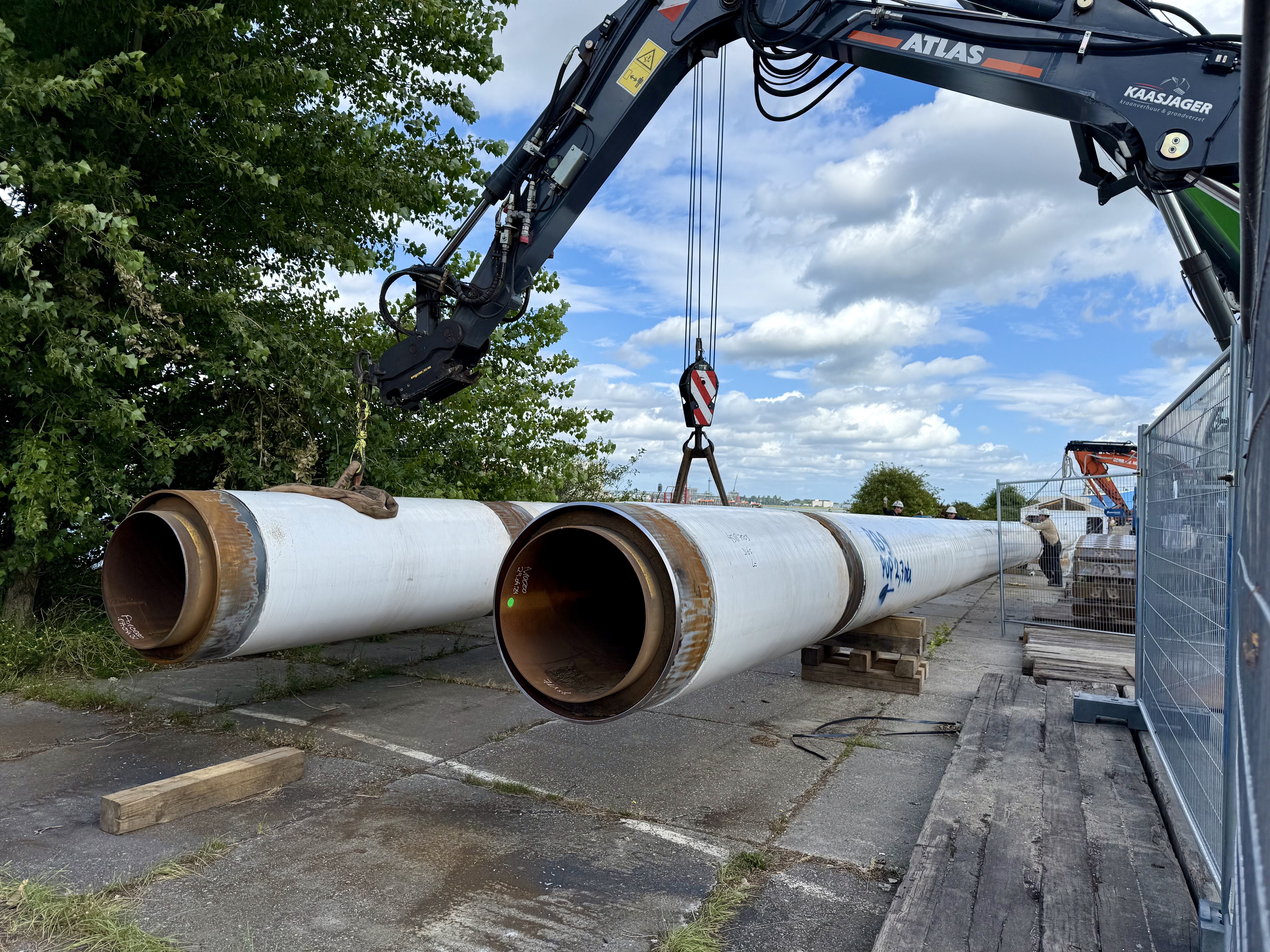 Een kraan verplaatst grote metalen leidingen van WarmtelinQ op een bouwplaats bij Vlaardingen
